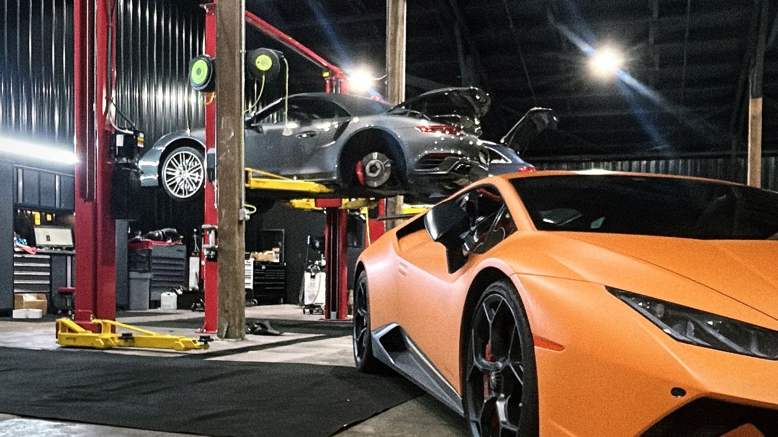 Orange Lamborghini Huracán beside a Porsche 911 on the lift at Bluegrass Precision Motorwerks
